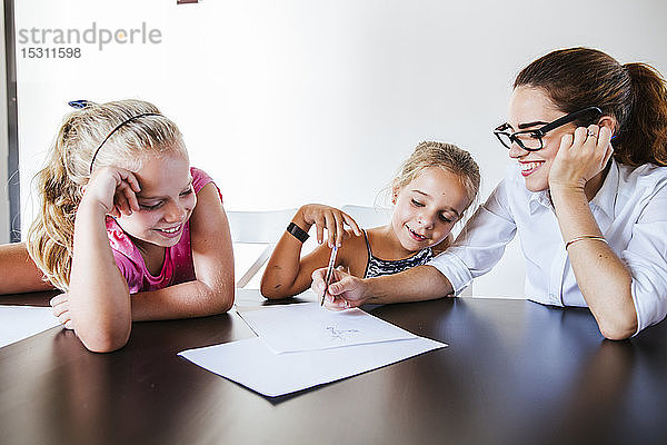 Glückliche Lehrerin sitzt am Schreibtisch mit zwei Schulmädchen  die auf Papier schreiben