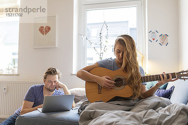 Junge Frau spielt zu Hause Gitarre mit beschädigter Saite mit Partner am Laptop