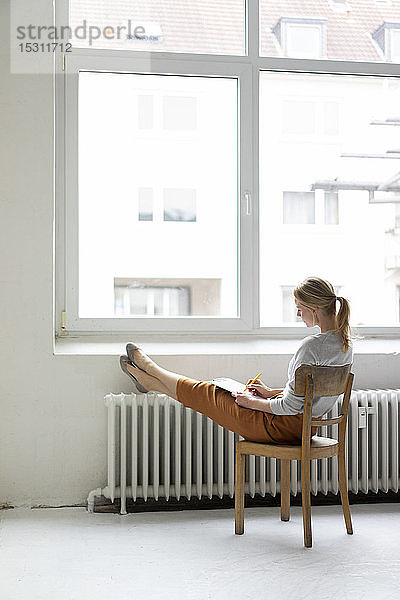 Junge Frau sitzt auf Stuhl am Fenster im Büro und macht Notizen
