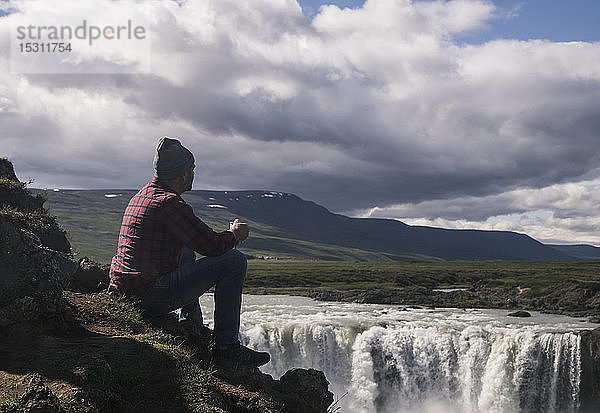 Mann sitzt mit einer Tasse Kaffee und schaut auf die Godafoss-Wasserfälle  Island
