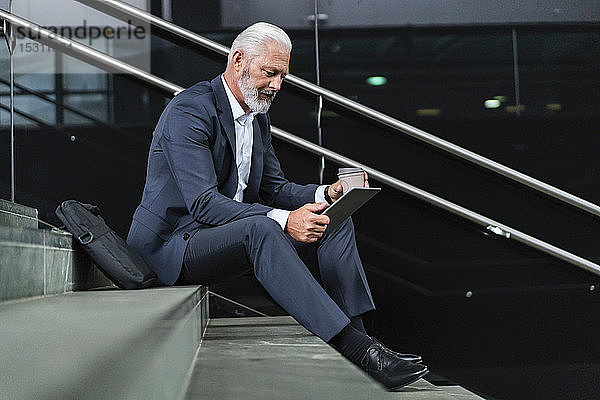 Reifer Geschäftsmann sitzt auf einer Treppe und benutzt ein Tablett