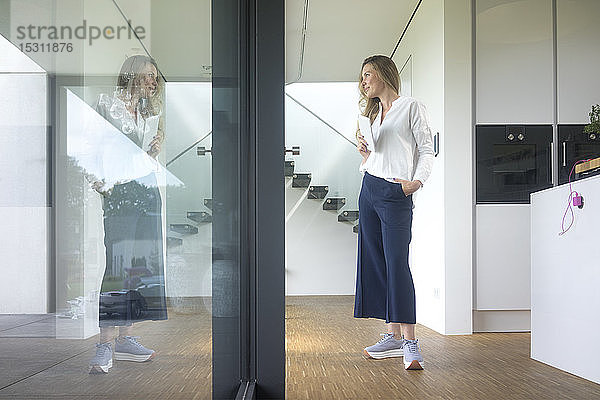 Geschäftsfrau hält Tablette in modernem Haus und schaut aus dem Fenster