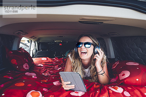 Hübsche blonde Frau mit Sonnenbrille und Kopfhörer beim Zelten in einem Lieferwagen mit Tablette