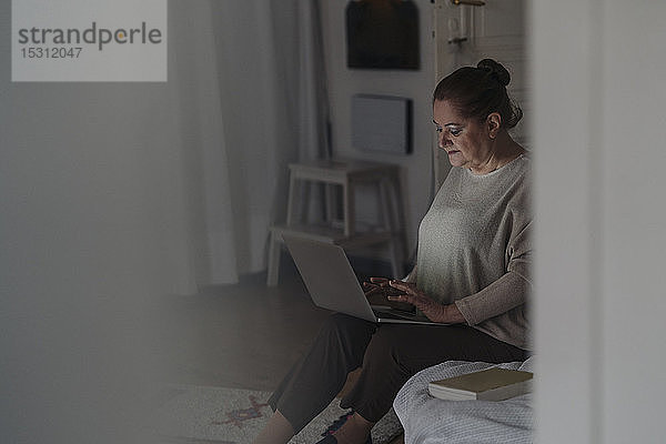 Ältere Frau sitzt zu Hause am Bett und benutzt einen Laptop