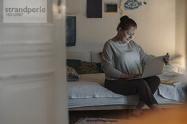 Ältere Frau sitzt zu Hause am Bett und benutzt einen Laptop