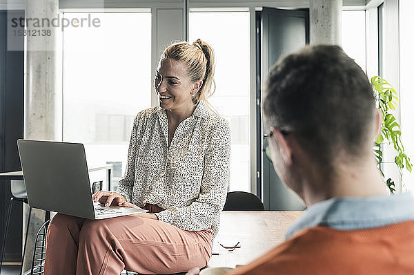 Lächelnde Geschäftsfrau sitzt im Büro mit Laptop auf dem Tisch  mit dem Geschäftsmann im Vordergrund