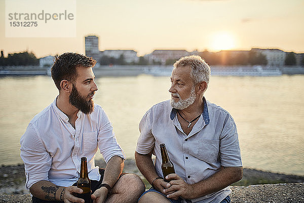 Vater und erwachsener Sohn sitzen bei Sonnenuntergang auf einer Mauer am Flussufer und trinken ein Bier