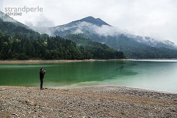 Mann steht am Seeufer und macht ein Foto mit seinem Smartphone  Stausee Wiestal  Bundesland Salzburg  Österreich