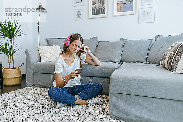 Lächelnde junge Frau sitzt auf dem Boden im Wohnzimmer und hört Musik mit ihrem Handy