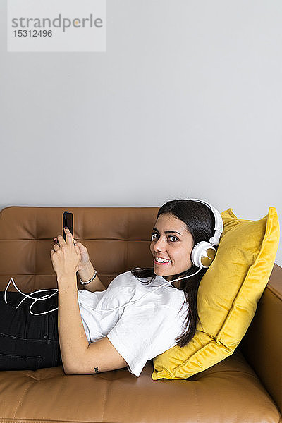 Junge Frau liegt zu Hause auf der Couch mit Smartphone und Kopfhörern