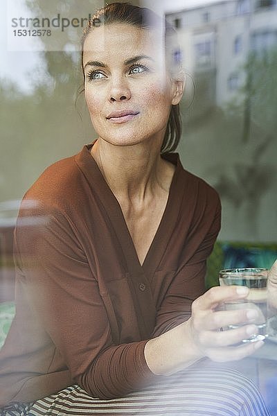 Porträt einer nachdenklichen Frau mit Getränk hinter einer Fensterscheibe