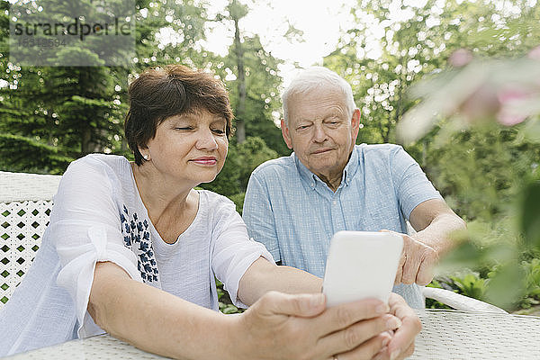 Porträt eines älteren Ehepaares  das am Gartentisch sitzt und auf sein Handy schaut