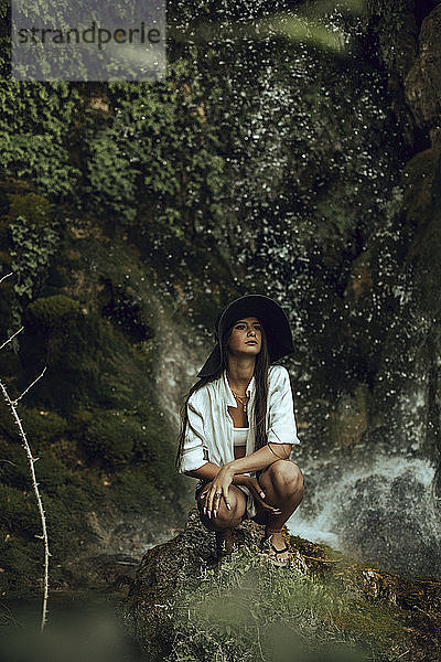 Junge Frau mit Hut  die in der Nähe eines Wasserfalls kauert