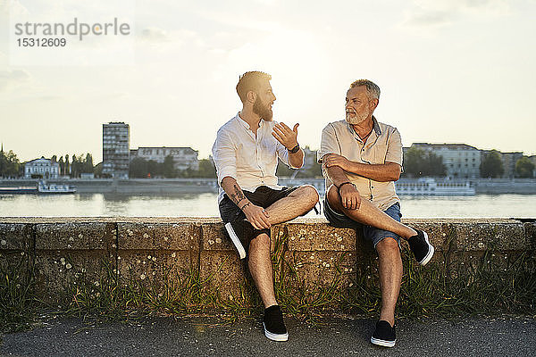 Vater und erwachsener Sohn sitzen auf einer Mauer am Flussufer und unterhalten sich