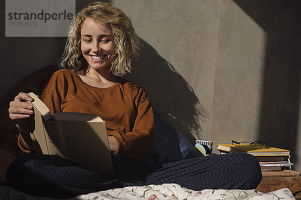 Porträt einer lächelnden jungen Frau  die im Bett sitzt und ein Buch liest