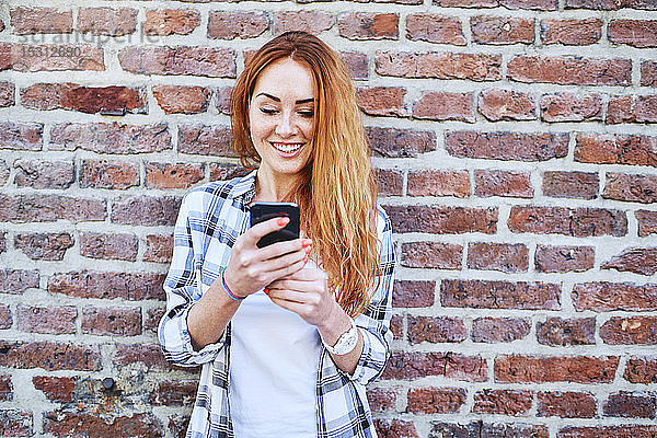 Fröhliche junge Frau benutzt Smartphone  während sie sich an eine Ziegelmauer lehnt