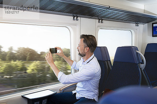 Älterer Mann sitzt in einem Zug und fotografiert mit seinem Smartphone