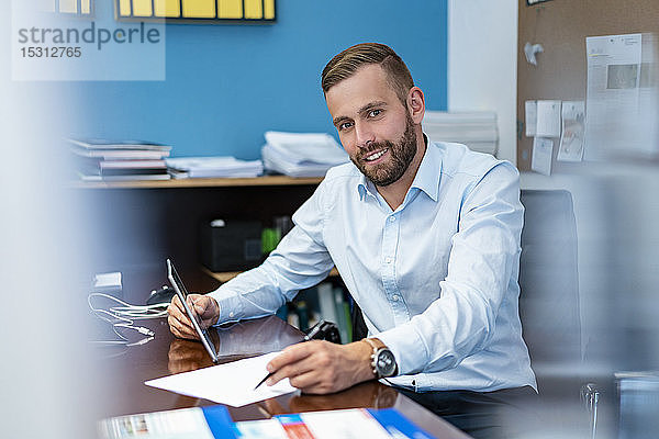 Porträt eines lächelnden Geschäftsmannes mit Papieren und Tablett am Schreibtisch im Büro
