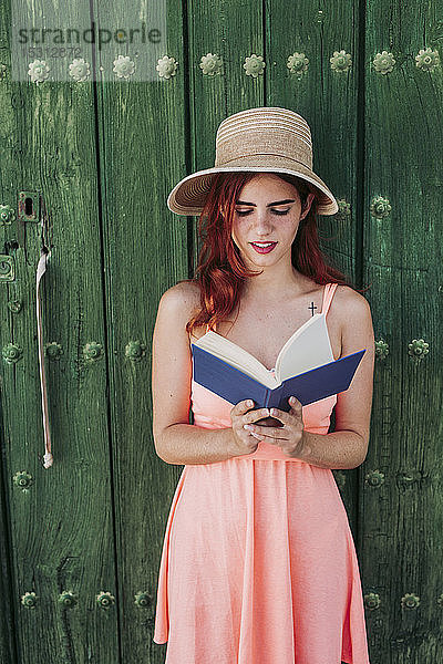 Porträt einer rothaarigen jungen Frau  die im Sommer vor einer grünen Holztür steht und ein Buch liest