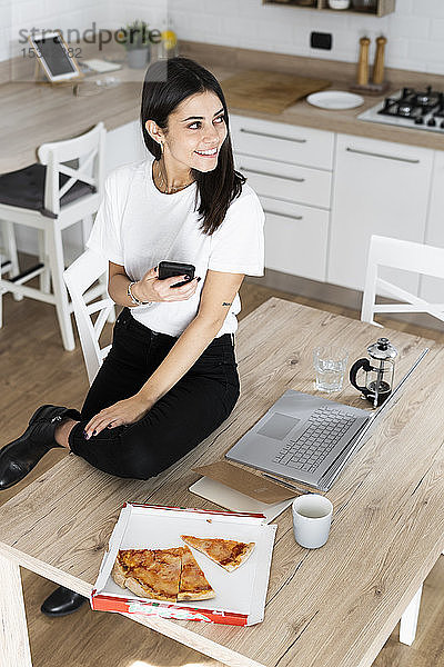 Junge Frau mit Handy beim Pizzaessen in der heimischen Küche