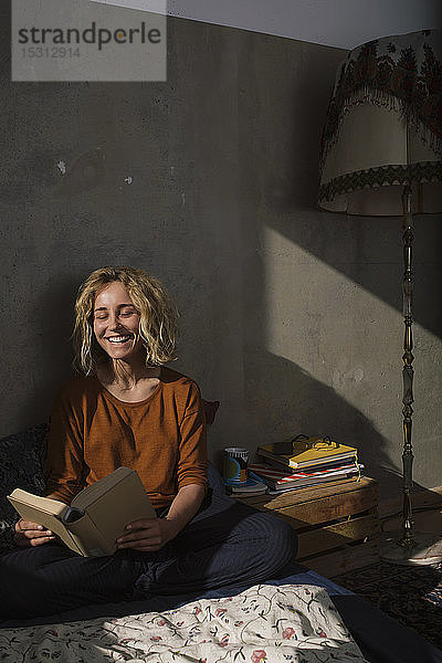 Porträt einer lachenden jungen Frau  die auf dem Bett sitzt und ein Buch liest