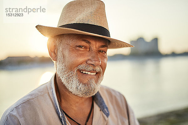 Porträt eines lächelnden älteren Mannes mit Sommerhut am Flussufer bei Sonnenuntergang
