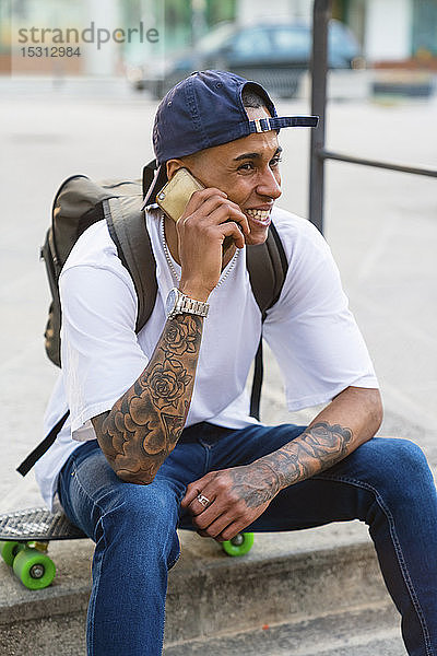 Porträt eines tätowierten jungen Mannes am Telefon  der im Freien auf seinem Skateboard sitzt