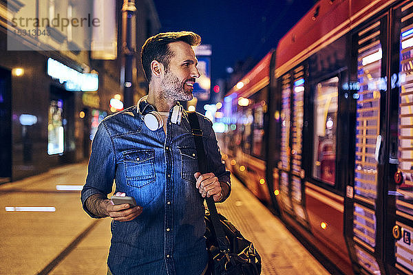Lächelnder Mann benutzt nachts in der Stadt sein Smartphone  während er auf die Straßenbahn wartet