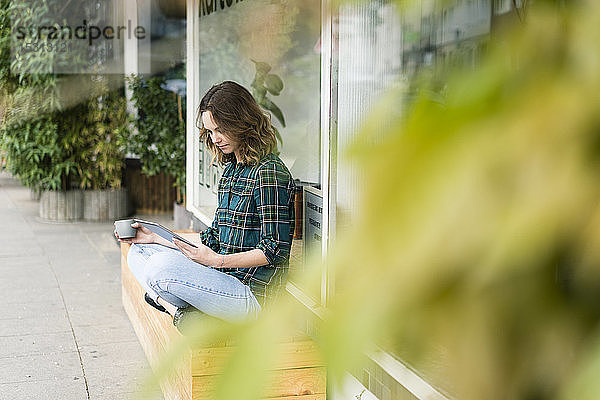 Junge Frau sitzt vor einem Café  benutzt ein digitales Tablet und trinkt Kaffee