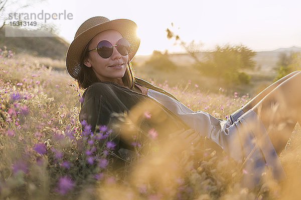 Junge Frau mit Sonnenbrille und Sonnenhut  die bei Sonnenuntergang auf einer Wiese sitzt