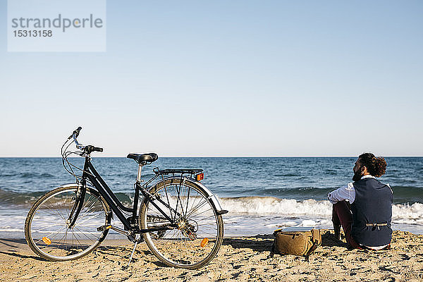 Gut gekleideter Mann mit seinem Fahrrad am Strand sitzend