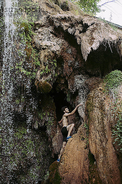 Barbusiger junger Mann klettert an einem Wasserfall