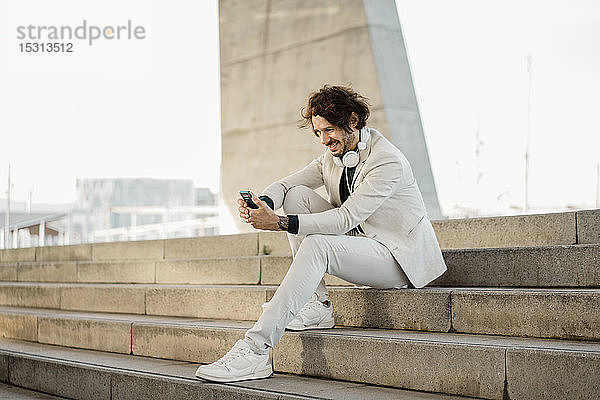 Lächelnder Geschäftsmann mit Kopfhörern sitzt auf einer Treppe im Freien und schaut auf sein Handy
