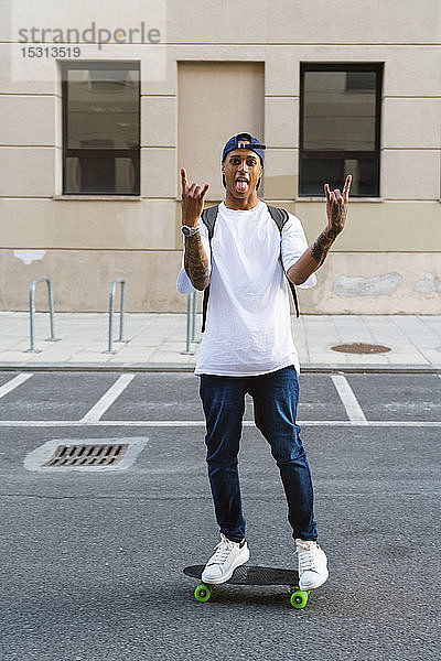 Tätowierter junger Mann steht auf Skateboard  streckt die Zunge heraus und zeigt Rock-and-Roll-Zeichen