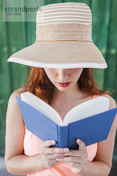 Rothaarige junge Frau vor grüner Holztür beim Lesen eines Buches im Sommer
