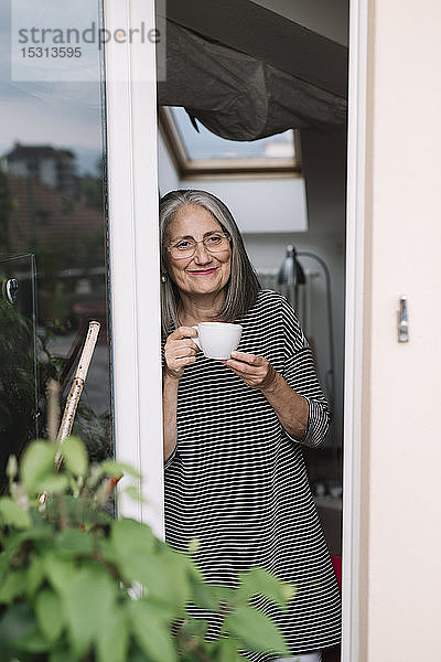 Porträt einer glücklichen älteren Frau  die sich an eine geöffnete Balkontür lehnt und mit einer Tasse Kaffee in die Ferne schaut