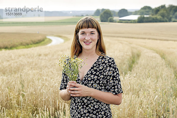 Porträt einer lächelnden jungen Frau mit Kamillenblütenstrauss vor einem Getreidefeld stehend