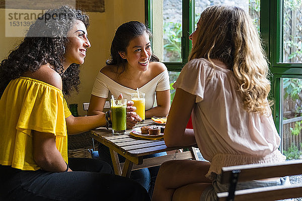 Drei glückliche junge Frauen mit Smoothies treffen sich in einem Cafe