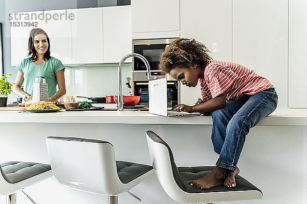 Mädchen sitzt mit Laptop auf dem Küchentisch mit der Mutter im Hintergrund