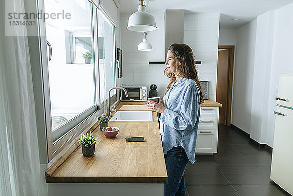 Junge Frau mit Pyjama in der Küche zu Hause  die aus dem Fenster schaut