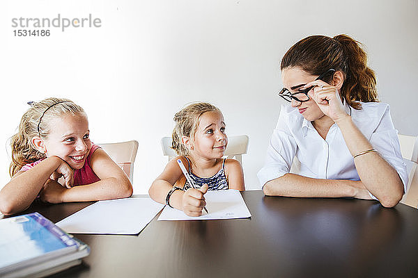Lehrerin sitzt am Schreibtisch mit zwei Schulmädchen  die auf Papier schreiben