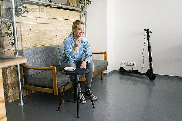 Junge Frau sitzt auf einer Couch in einem Kaffeehaus  im Hintergrund lädt ein E-Scooter auf