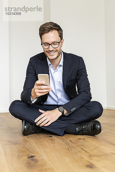 Lächelnder Geschäftsmann sitzt auf dem Boden und benutzt sein Handy