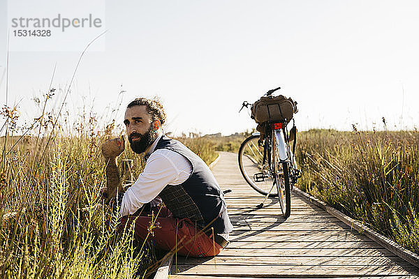 Gut gekleideter Mann  der auf einem Holzsteg auf dem Land neben einem Fahrrad sitzt