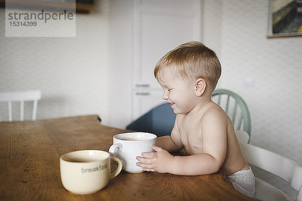 Lachender kleiner Junge sitzt am Küchentisch und hält eine Tasse