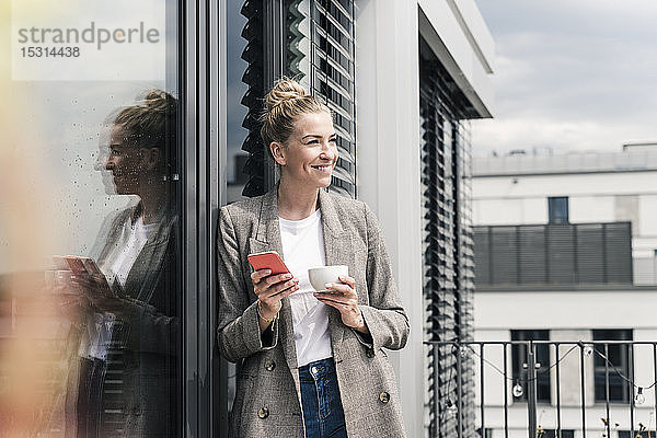 Lächelnde Geschäftsfrau mit Handy und Kaffeetasse auf der Dachterrasse stehend