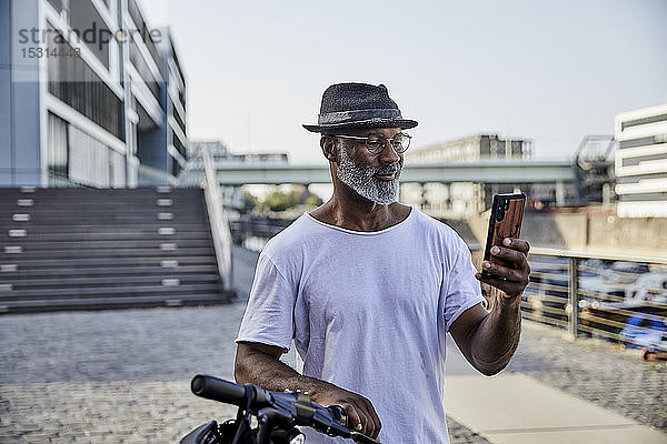 Porträt eines lächelnden reifen Mannes mit E-Scooter  der auf ein Mobiltelefon schaut