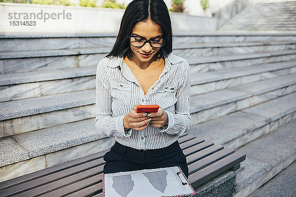 Junge Geschäftsfrau  die auf einer Bank sitzt und ihr Handy überprüft