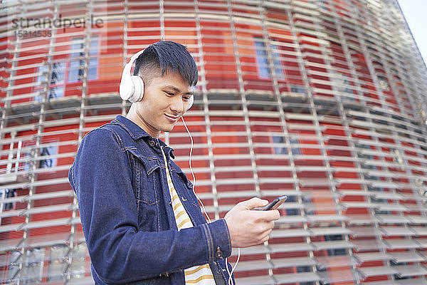 Porträt eines lächelnden jungen Mannes mit Kopfhörern  der auf sein Handy schaut  Barcelona  Spanien