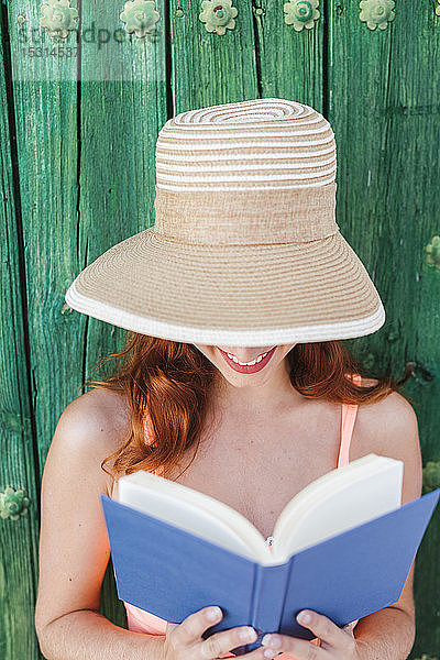 Lächelnde junge Frau vor grüner Holztür beim Lesen eines Buches